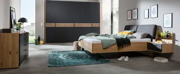 Schlafzimmer mit Holzbett, grauem Polsterkopfteil, schwarzem Kleiderschrank mit Holzleisten und Nachttisch rechts.